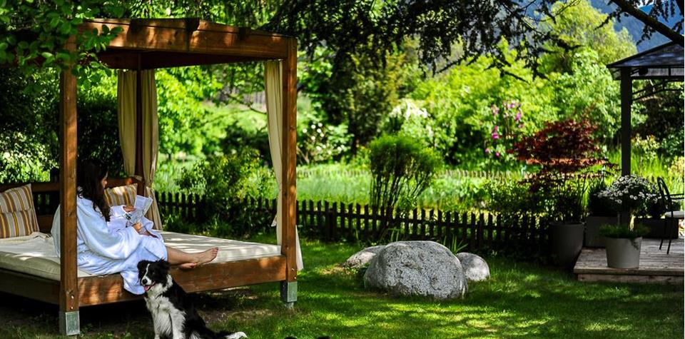 A woman is sitting on a covered bed in the garden, with two dogs lying on the grass in front of her