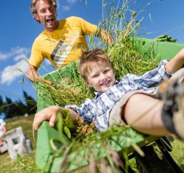 A man is pushing a wheelbarrow, and a child is sitting inside it