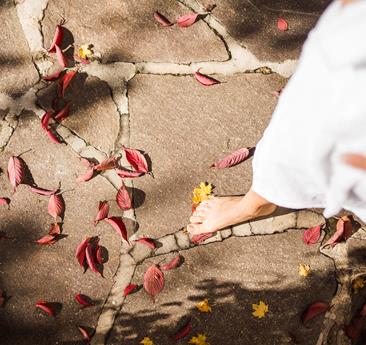 A person in a white bathrobe walks barefoot along a stone path covered with fallen leaves