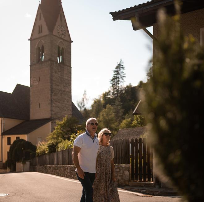 Ein Mann und eine Frau gehen eine ruhige Straße entlang, im Hintergrund erhebt sich eine Kirche