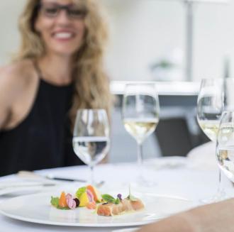 A woman sits smiling at an elegantly set table, enjoying a glass of white wine