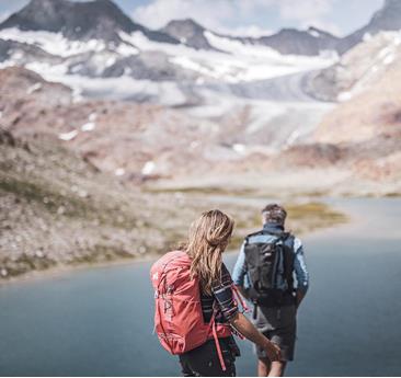 A couple is hiking along the shore of a mountain lake, surrounded by glaciers and alpine peaks