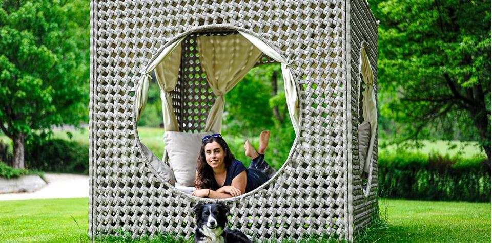 A woman is lying in a woven outdoor lounge while a dog is resting on the grass in front