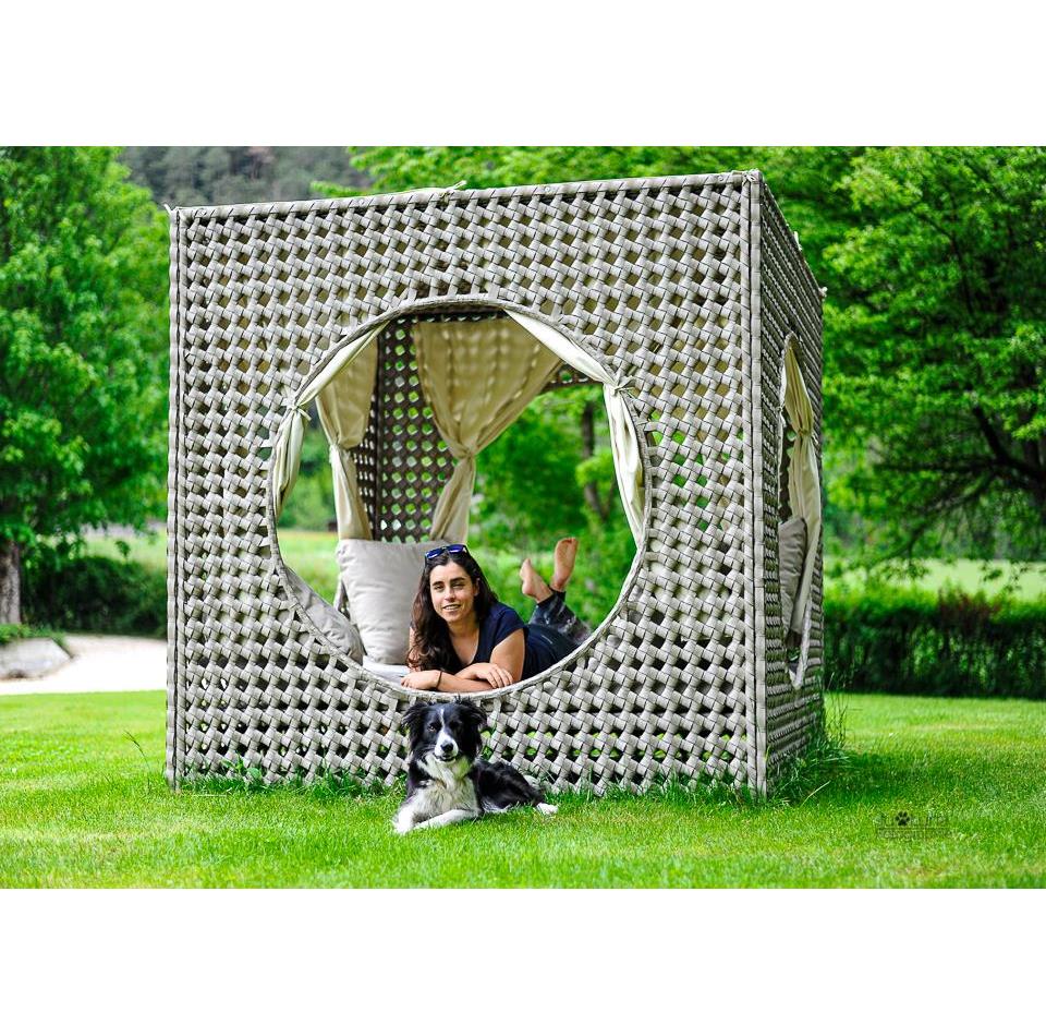 A woman is lying in a woven outdoor lounge while a dog is resting on the grass in front