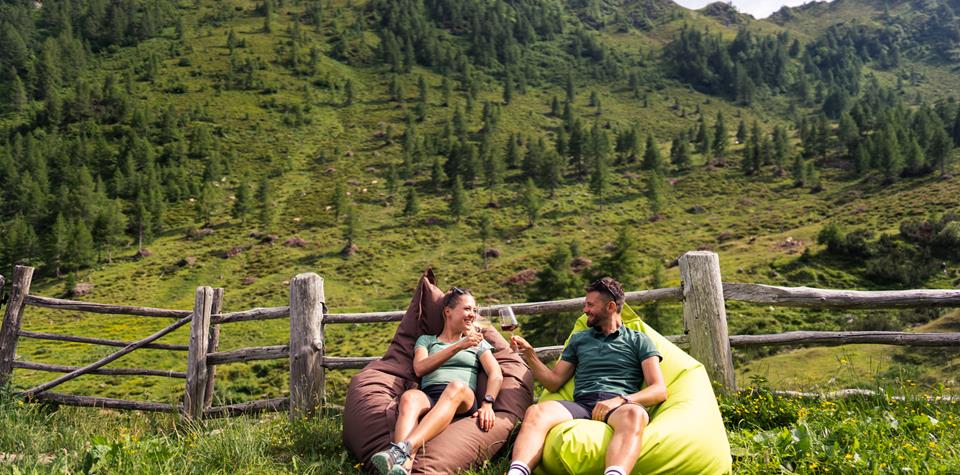 A couple is sitting relaxed on beanbags in front of an alpine meadow, toasting with glasses of wine