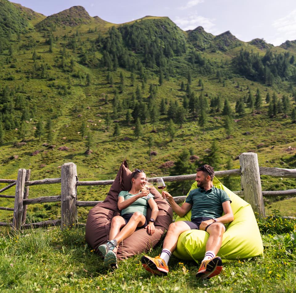 A couple is sitting relaxed on beanbags in front of an alpine meadow, toasting with glasses of wine
