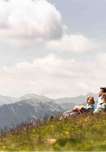 Eine Familie sitzt entspannt auf einer Bergwiese und genießt den Ausblick auf die umliegenden Berge